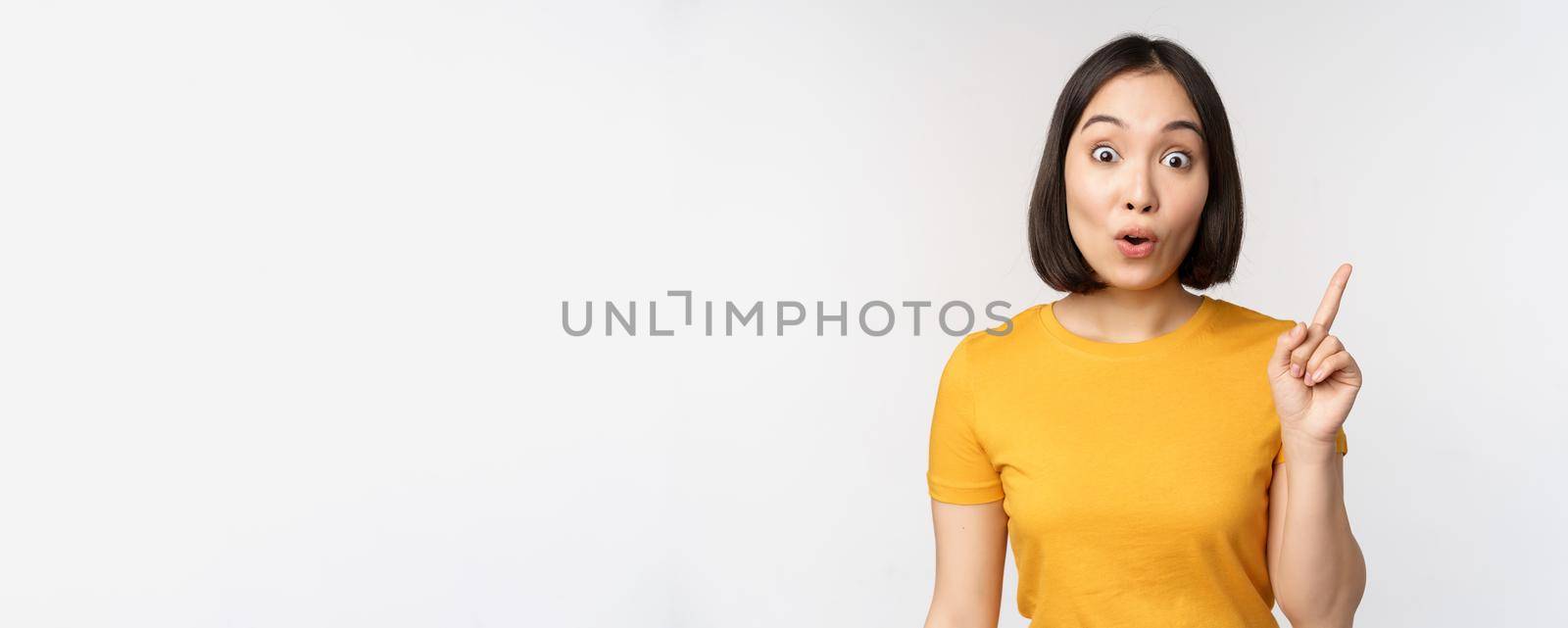 Beautiful young asian woman pointing finger up, smiling and looking amused at camera, showing advertisement, announcement on top, white background.