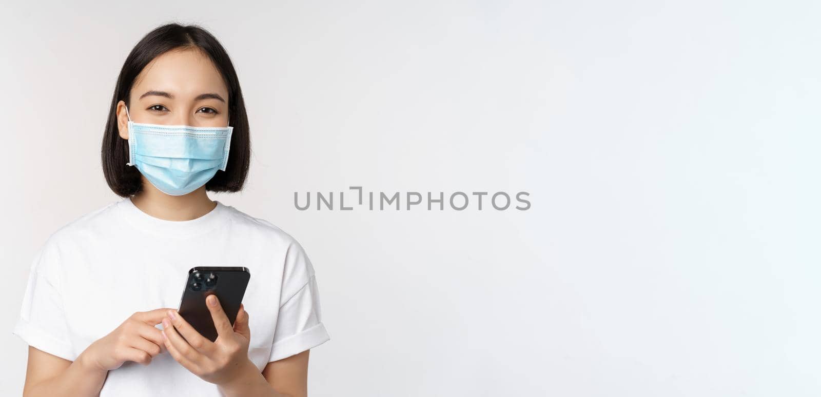 Health and covid-19 concept. Asian girl in medical face mask, using mobile phone application on quarantine, standing over white background by Benzoix