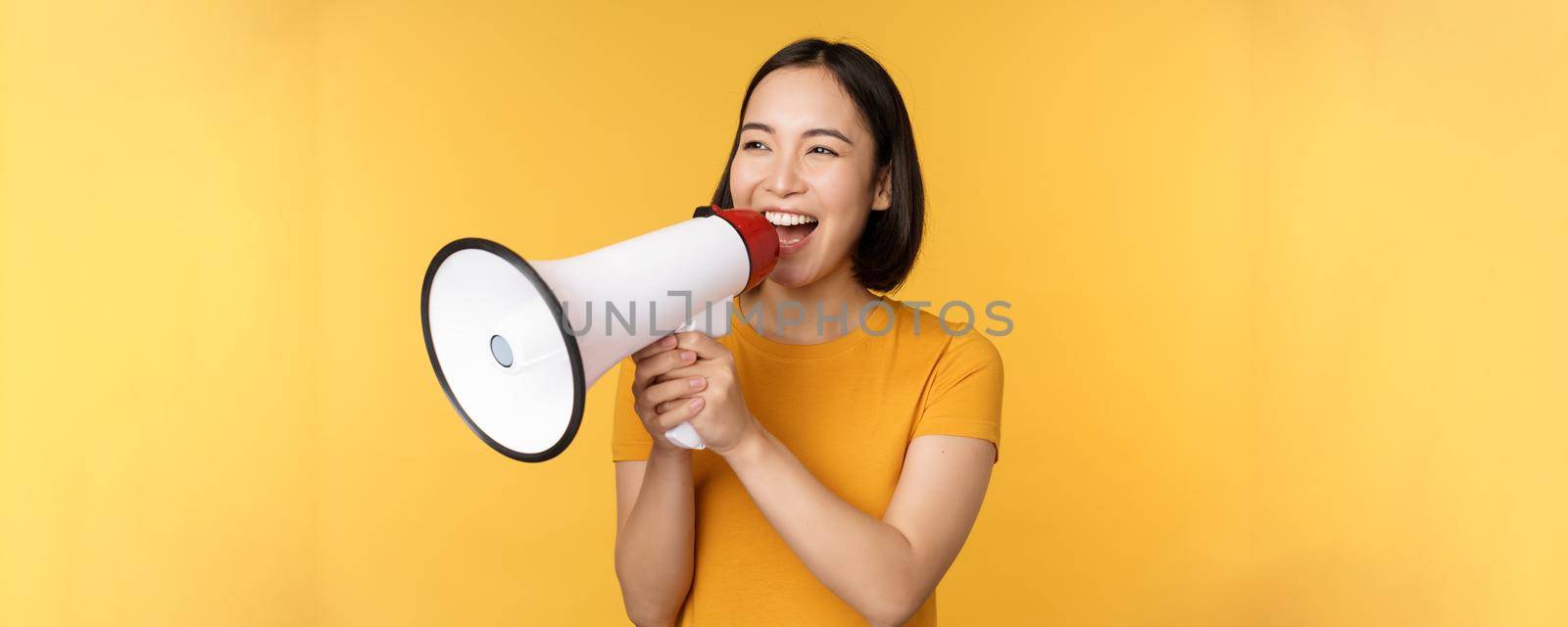 Announcement. Happy asian woman shouting loud at megaphone, recruiting, protesting with speaker in hands, standing over yellow background by Benzoix