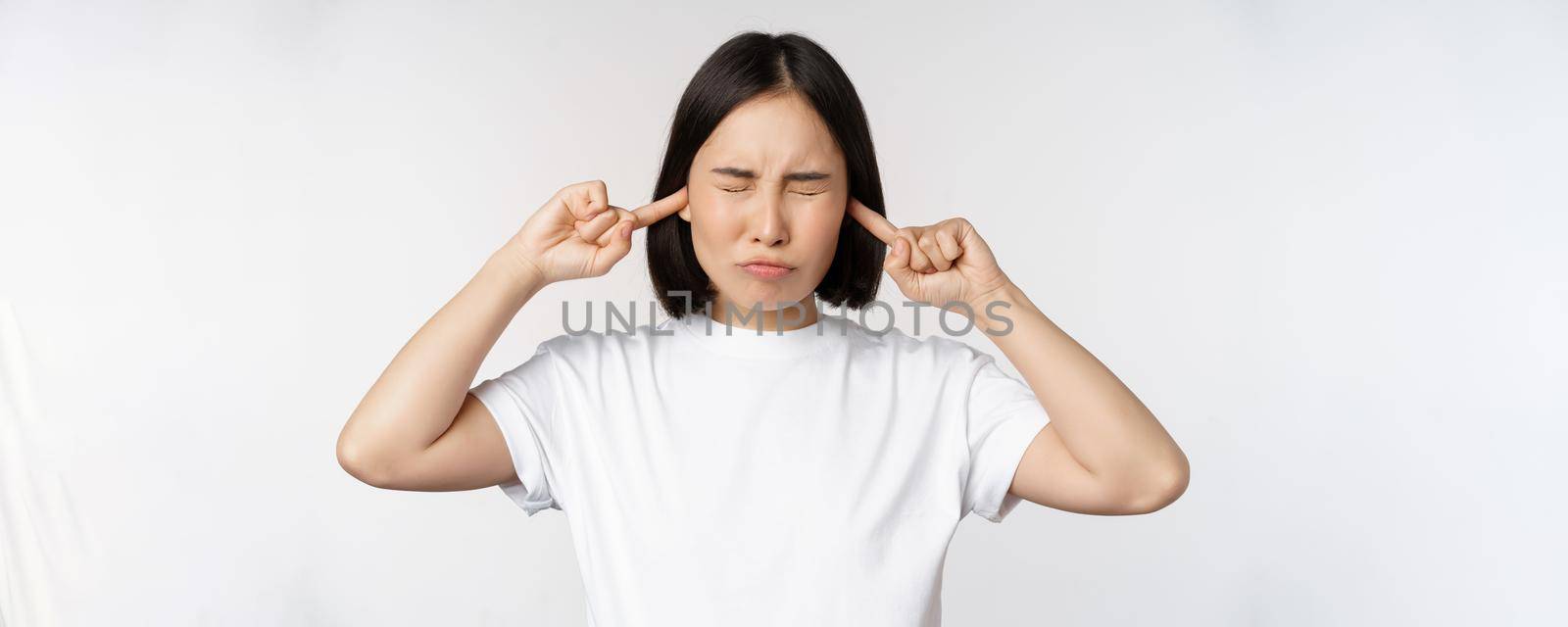 Portrait of asian woman shut ears and feeling discomfort from loud noise, annoying sound, standing over white background by Benzoix