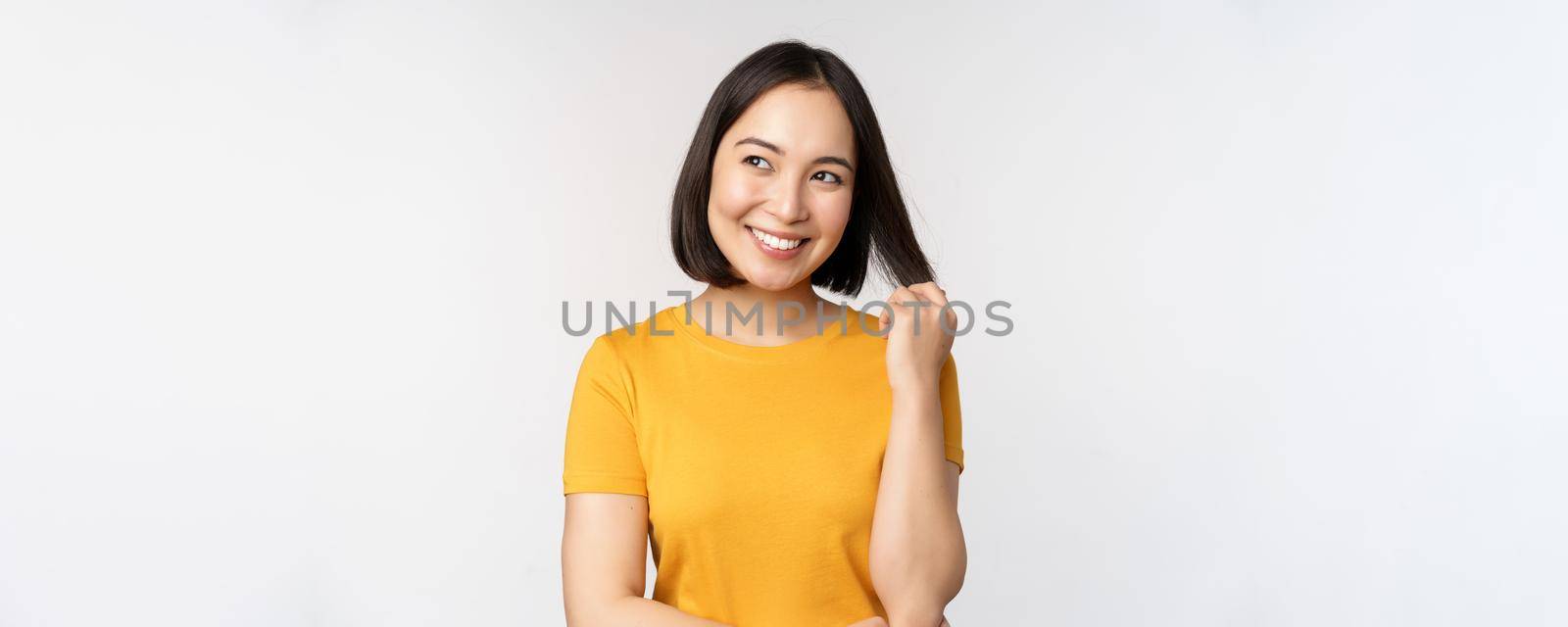 Beautiful romantic asian girl, smiling and playing with hair, looking happy at camera, standing in yellow t-shirt over white background.