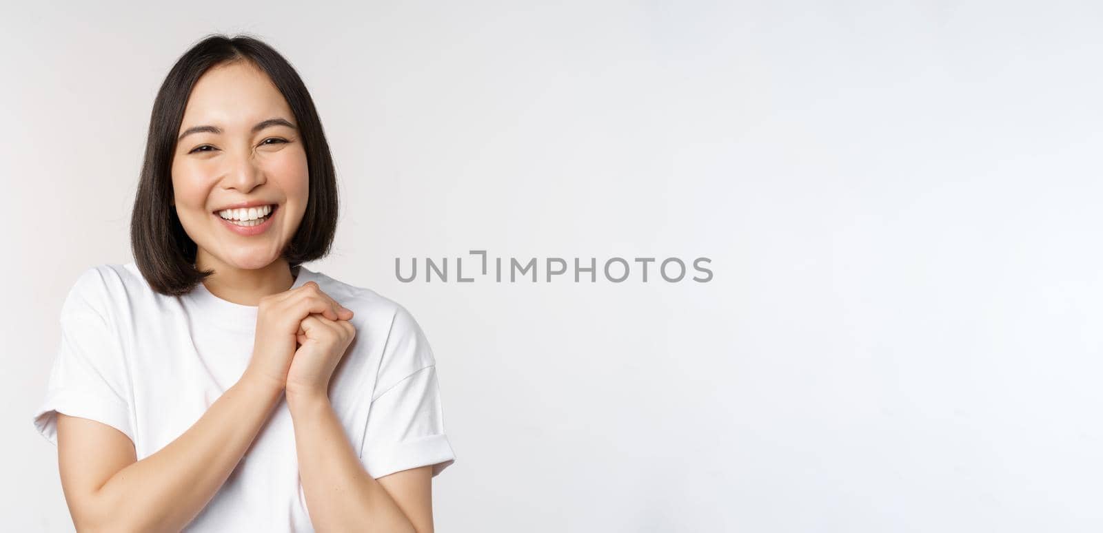Portrait of beautiful korean woman with healthy white smile, laughing and looking happy at camera, standing in tshirt over white studio background by Benzoix