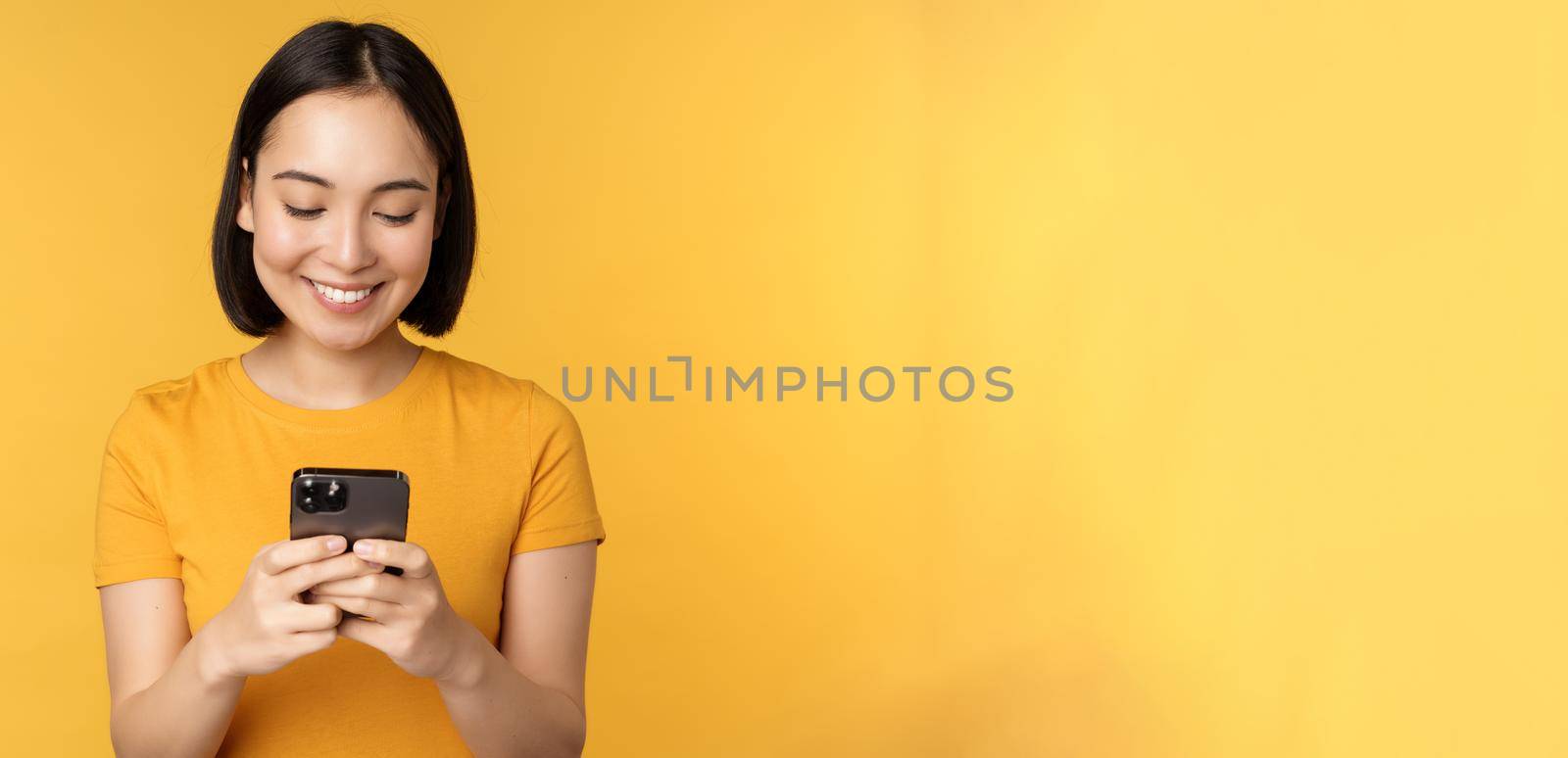 Technology. Smiling asian woman using mobile phone, holding smartphone in hands, standing in t-shirt against yellow background.