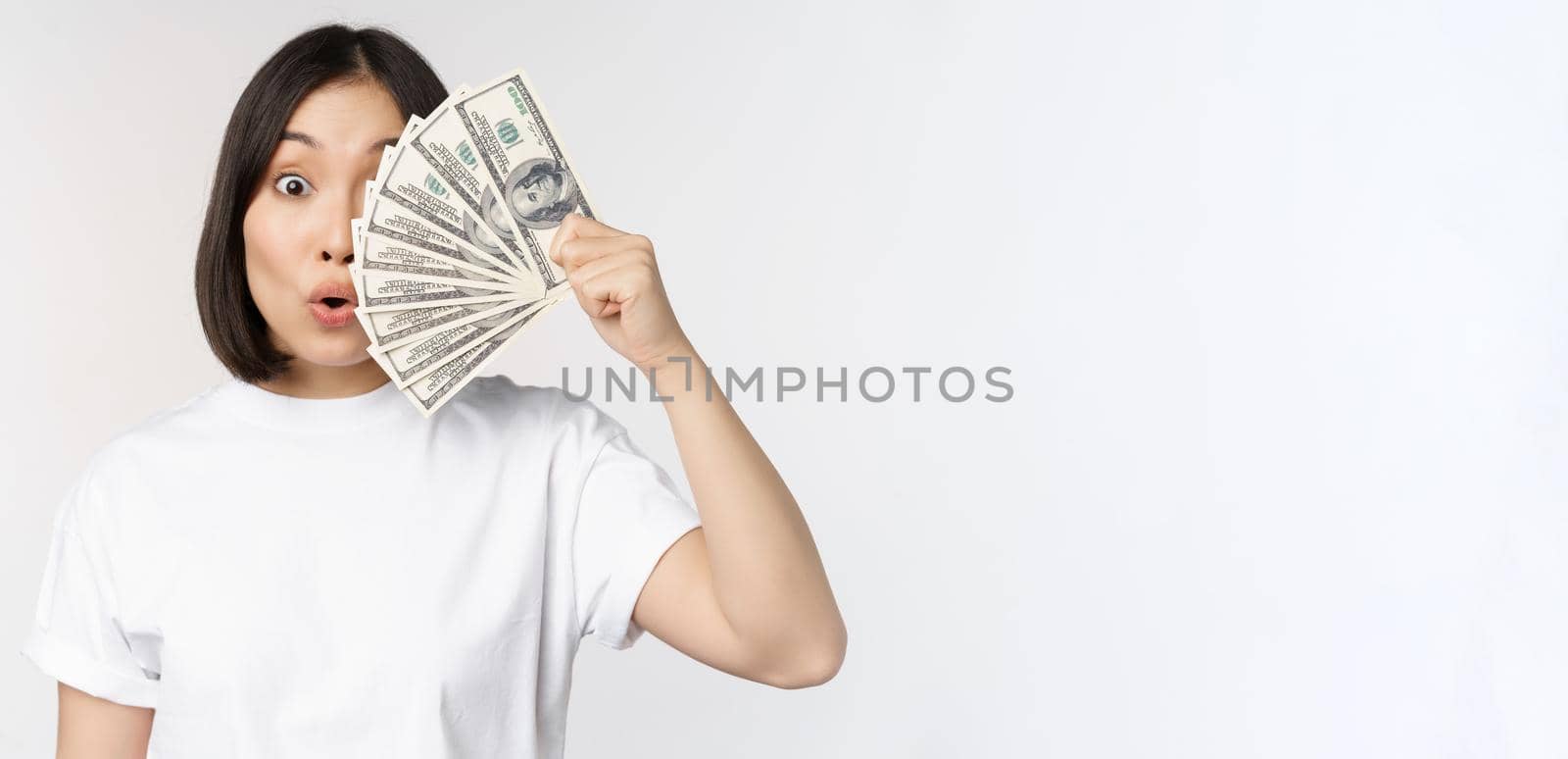 Happy asian girl holding money, cash near face, concept of microcredit and finance, white studio background by Benzoix