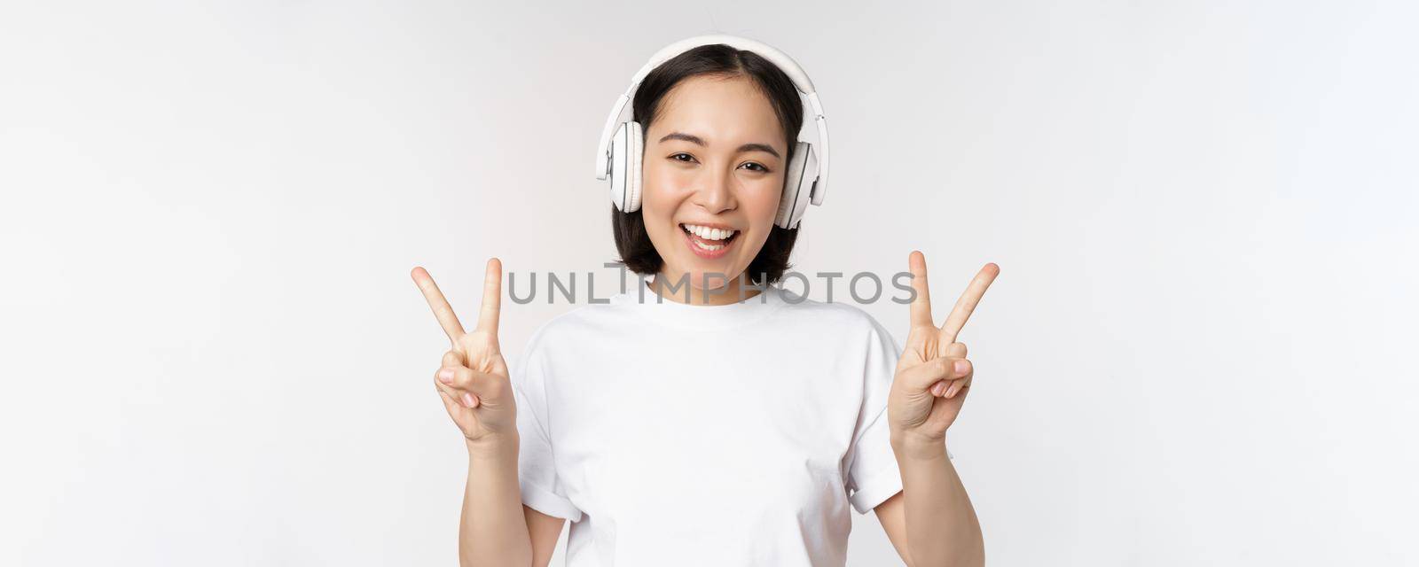 Happy asian woman wearing headphones and smiling, showing peace v-sign, listening music, standing in tshirt over white background by Benzoix