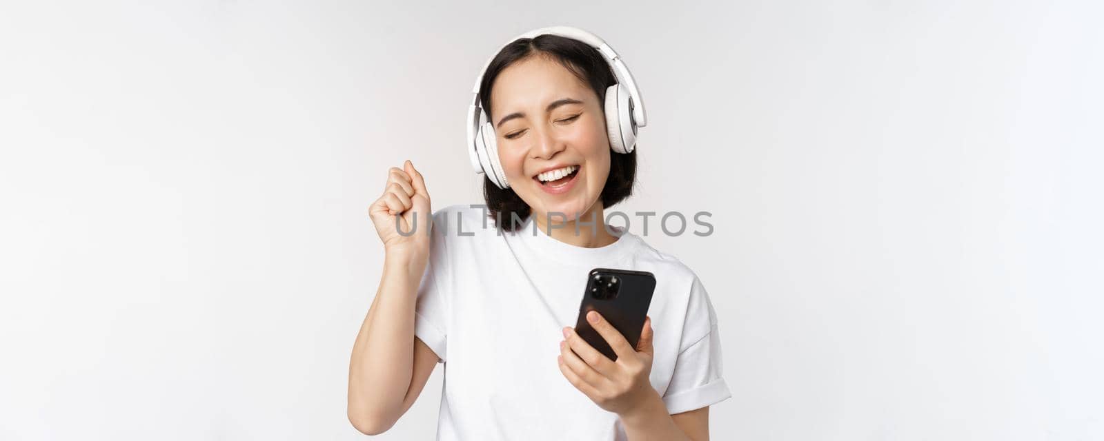 Beautiful modern asian girl, listening music in headphones, holding mobile phone, using smartphone app, standing over white background.