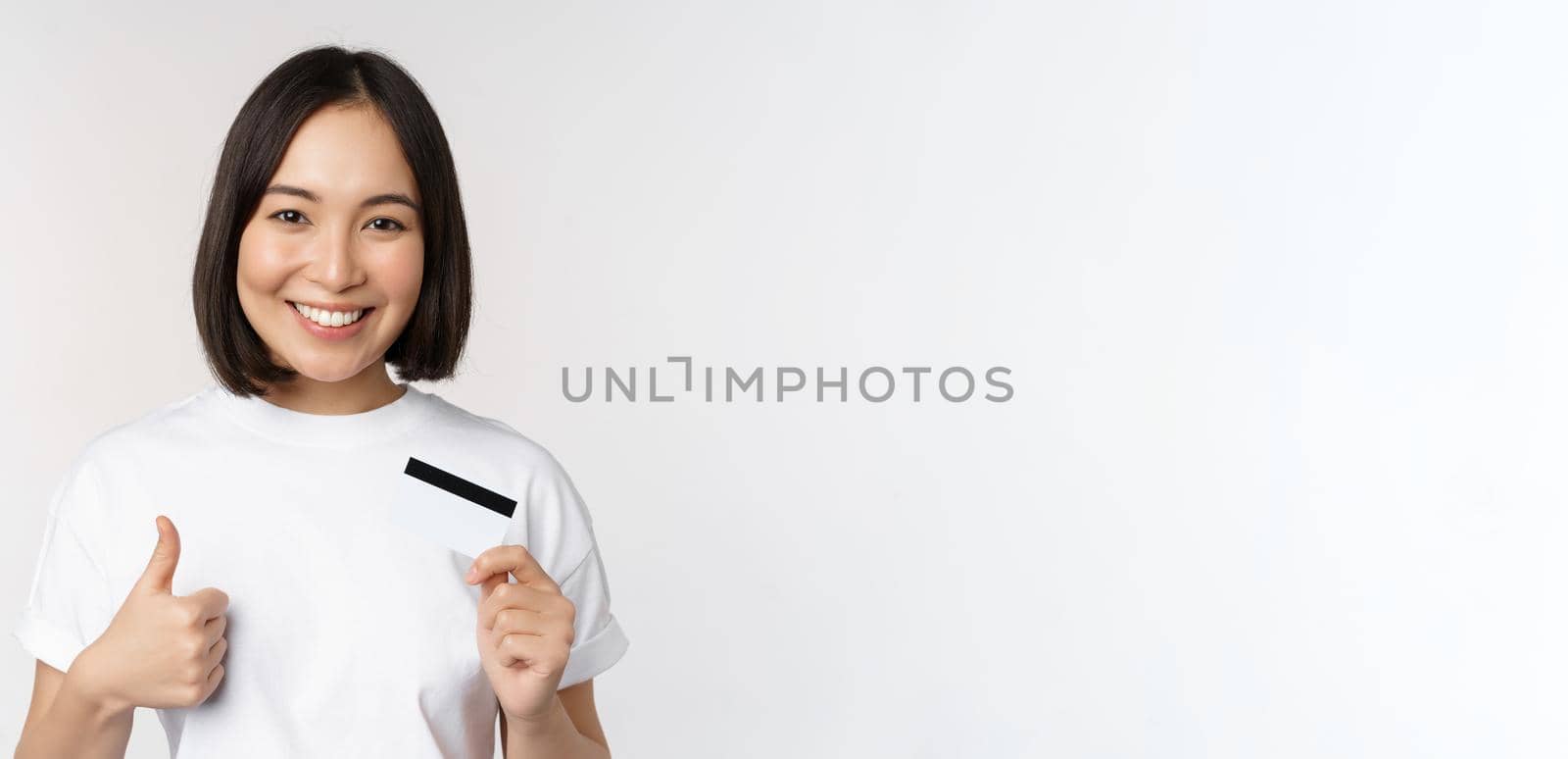 Portrait of beautiful young modern asian woman, showing credit card and thumbs up, recommending contactless payment, standing over white background.