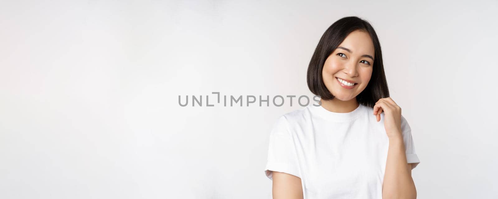 Portrait of cute coquettish woman laughing and smiling, looking aside thoughtful, thinking or imaging smth, standing in white t-shirt over studio background by Benzoix