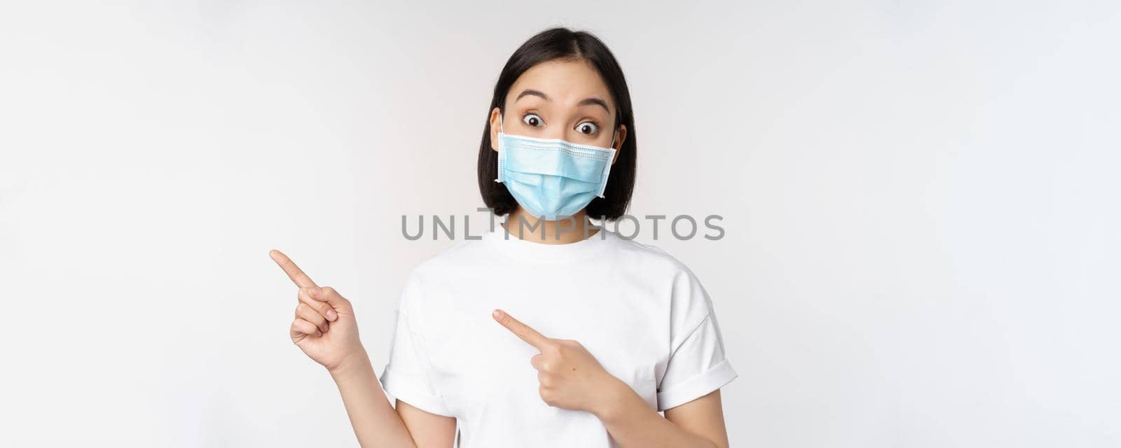 Surprised asian girl in medical mask, pointing fingers left, showing promo offer, raising eyebrows, amazed reaction, standing against white background.