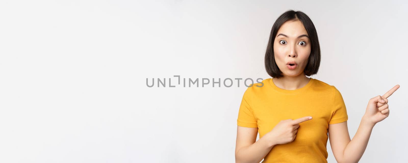 Amazed young asian woman, showing advertisement aside, pointing fingers right at promotion text, brand logo, standing happy against white background by Benzoix