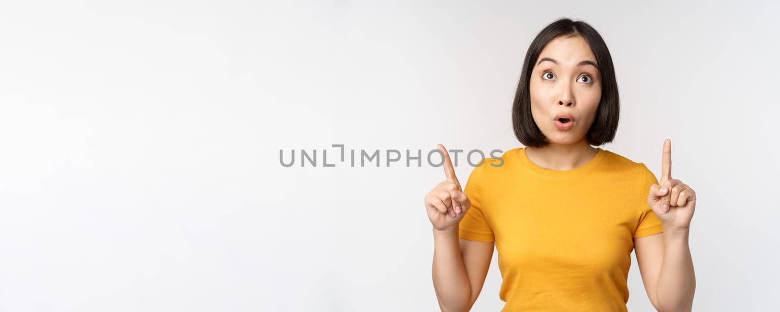 Happy beautiful asian girl showing advertisement, pointing fingers up, standing over white background.