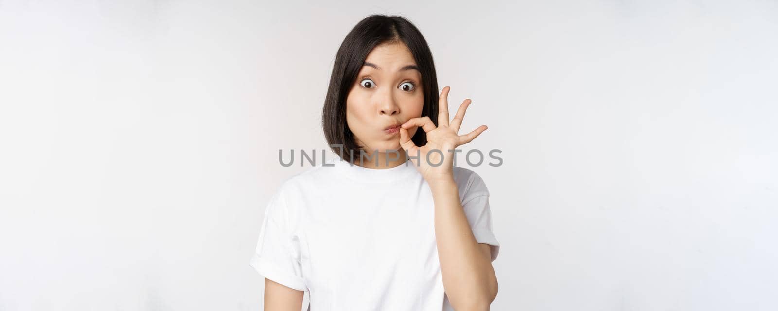 Image of young asian woman showing silence gesture, zipping mouth, seal lips, make promise, standing over white background by Benzoix