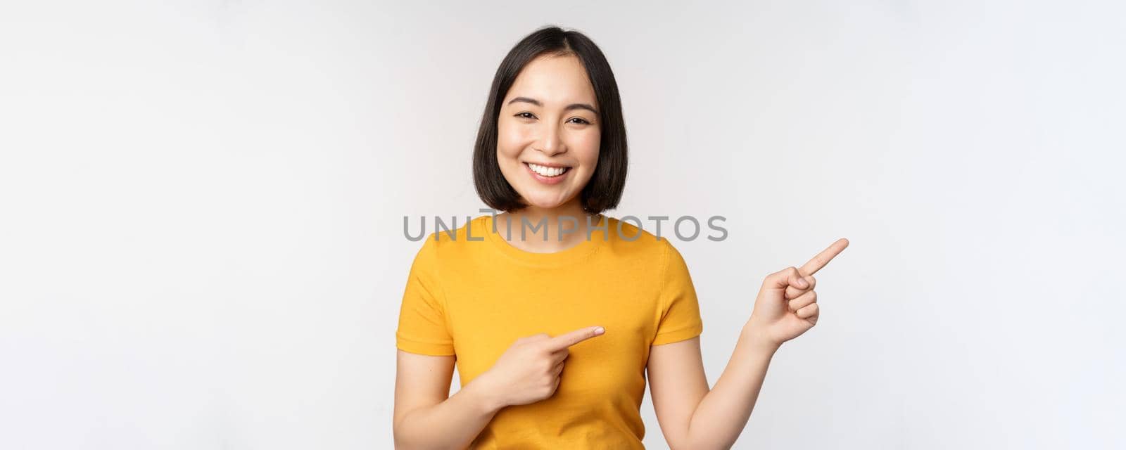 Amazed young asian woman, showing advertisement aside, pointing fingers right at promotion text, brand logo, standing happy against white background by Benzoix