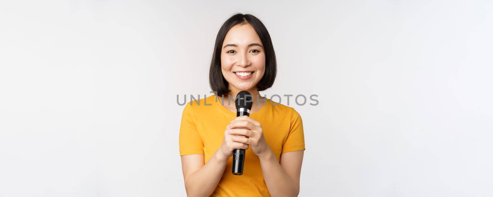 Image of young asian woman talking in microphone, perfom with mic, giving speech, standing in yellow tshirt against white background.