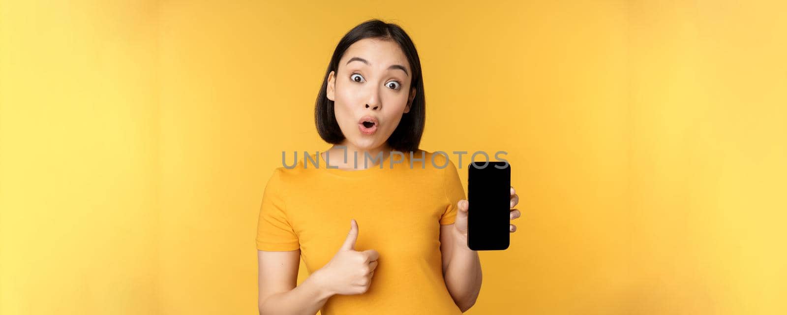 Excited asian woman showing mobile phone screen, thumbs up, like smth good, recommending smartphone application, standing over yellow background by Benzoix