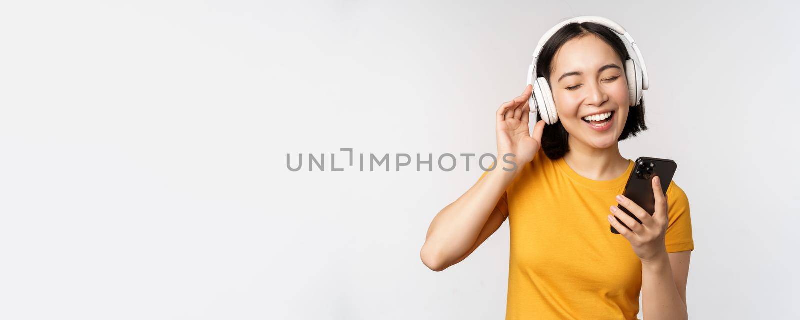 Cute japanese girl in headphones, looking at mobile phone and smiling, using music app on smartphone, standing against white background.