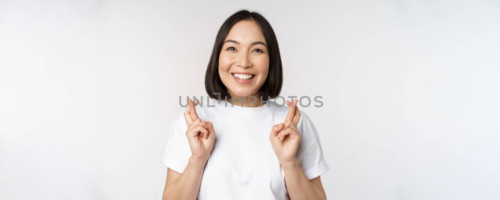 Hopeful asian girl cross fingers, making wish, anticipating, wishing smth, standing in tshirt over white background by Benzoix