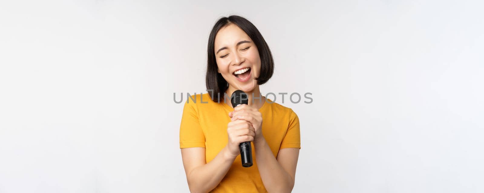 Happy asian girl singing and having fun, holding microphone at karaoke, standing in yellow tshirt against white background by Benzoix