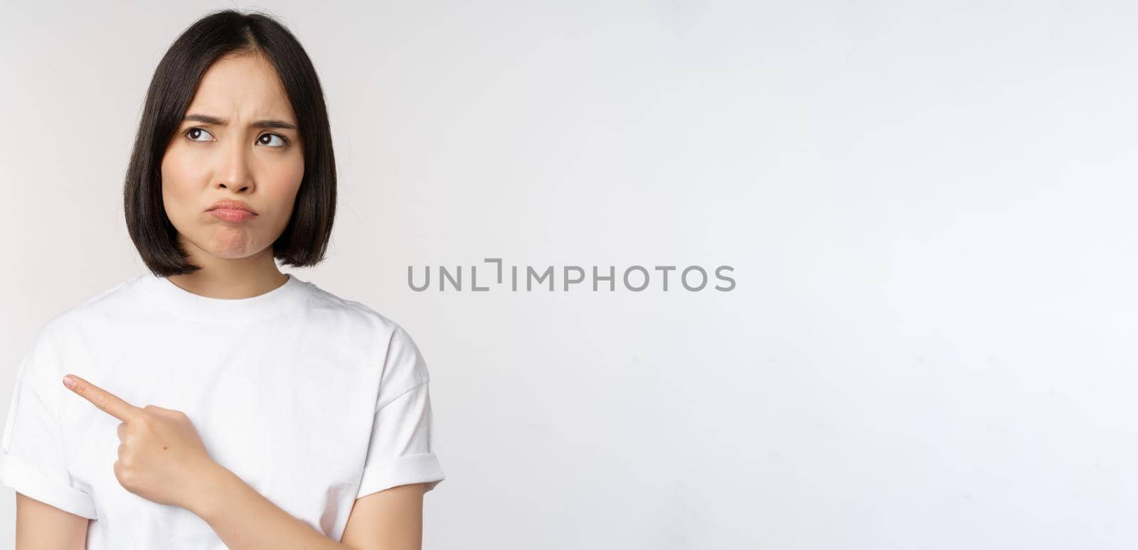 Angry and upset young asian woman pointing left, looking with regret or jealousy, feel unfair, standing over white background by Benzoix
