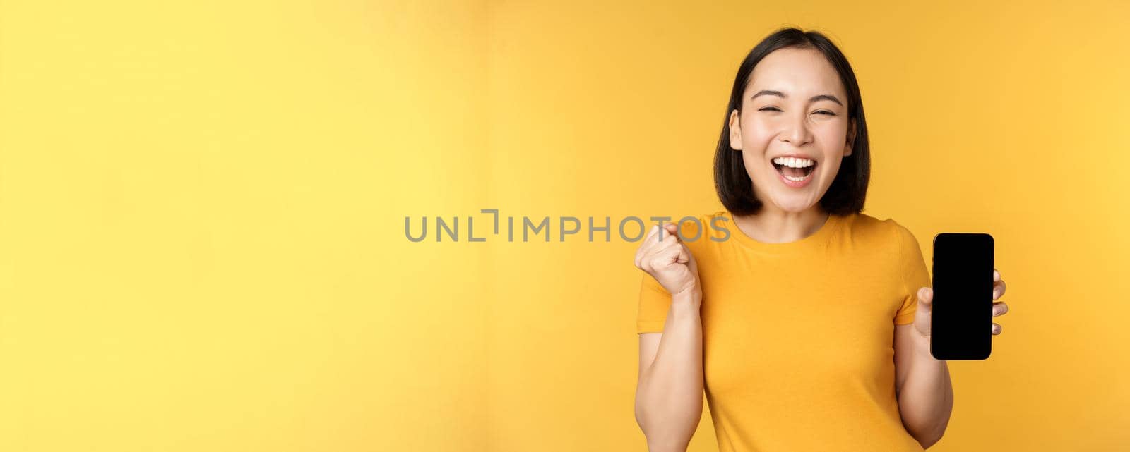 Excited asian woman, showing smartphone app and triumphing, celebrating on mobile phone, standing over yellow background by Benzoix