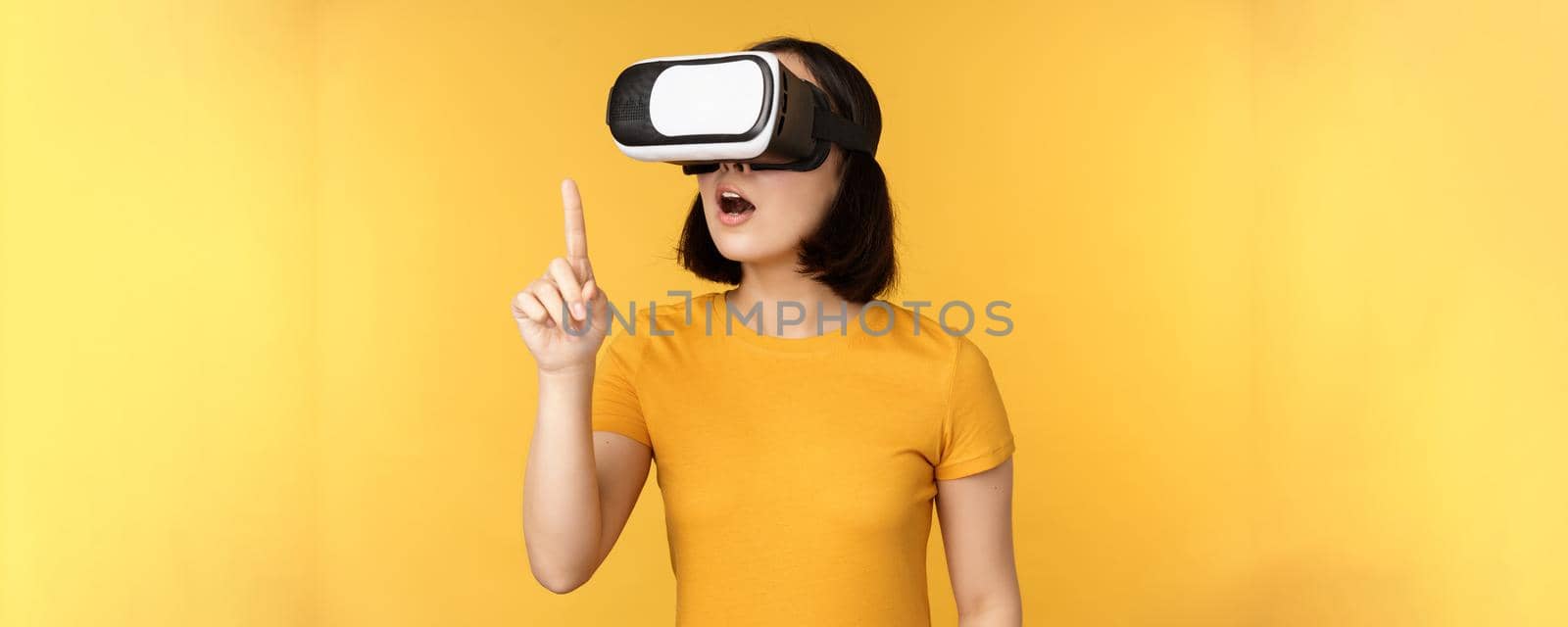 Girl in VR. Beautiful young asian woman, using virtual reality glasses and playing, chatting virtually, standing over yellow background by Benzoix