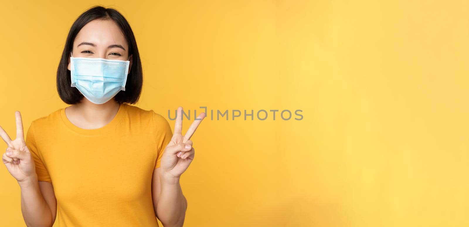 Positive asian woman smiling, wearing medical face mask from covid-19 during pandemic, showing peace v-sign gesture, yellow background.