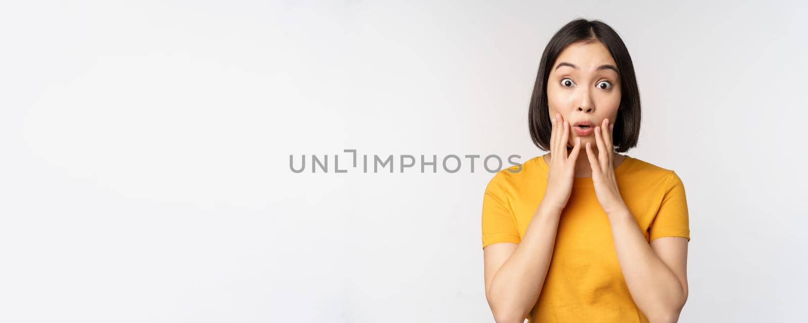 Close up portrait of asian woman looking surprised, wow face, staring impressed at camera, standing over white background in yellow t-shirt.
