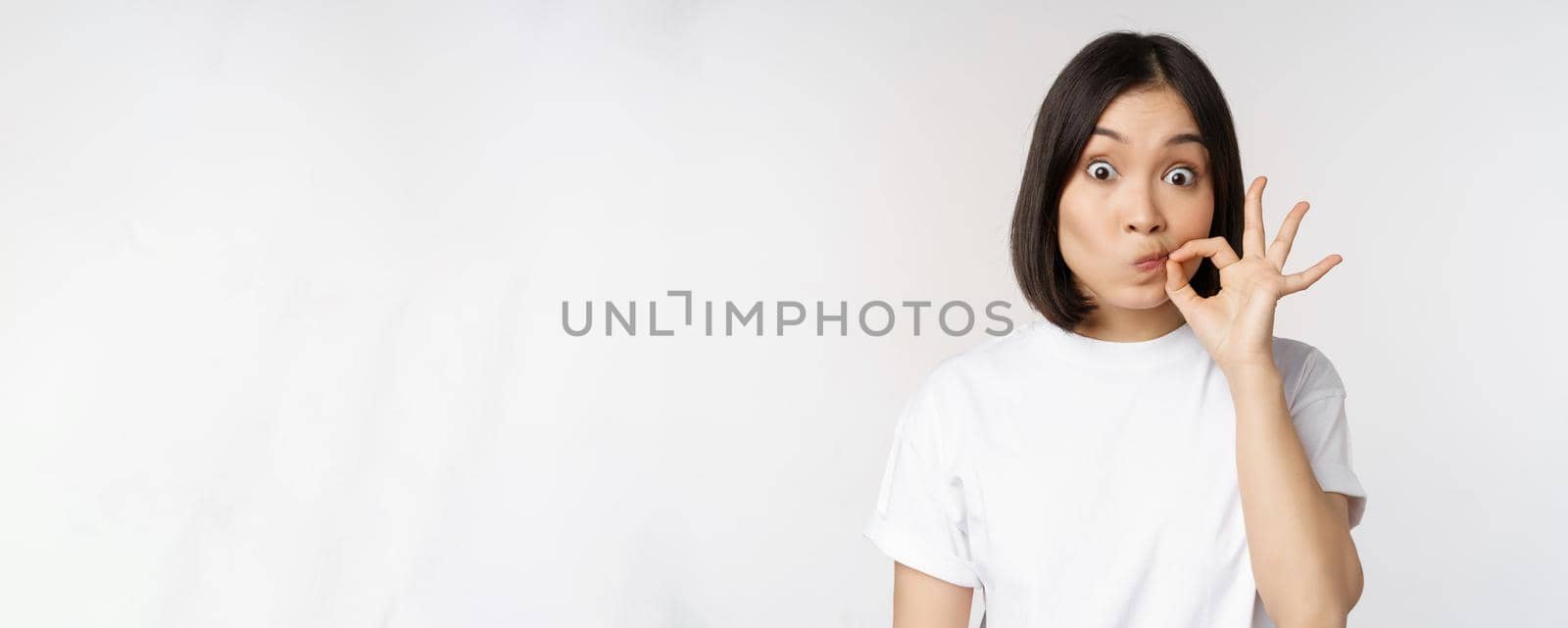 Image of young asian woman showing silence gesture, zipping mouth, seal lips, make promise, standing over white background by Benzoix