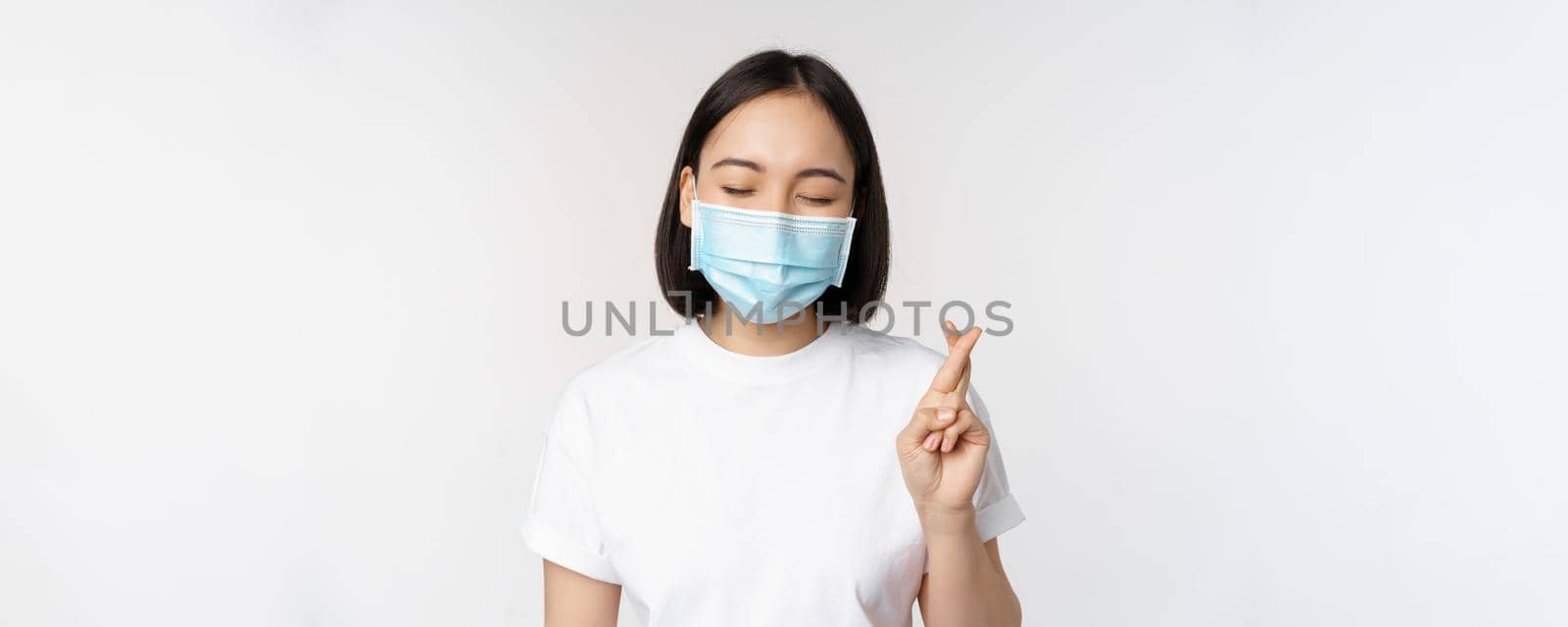 Covid-19, healthcare and medical concept. Image of asian girl in medical face mask, cross fingers, praying, making wish and smiling, standing over white background by Benzoix