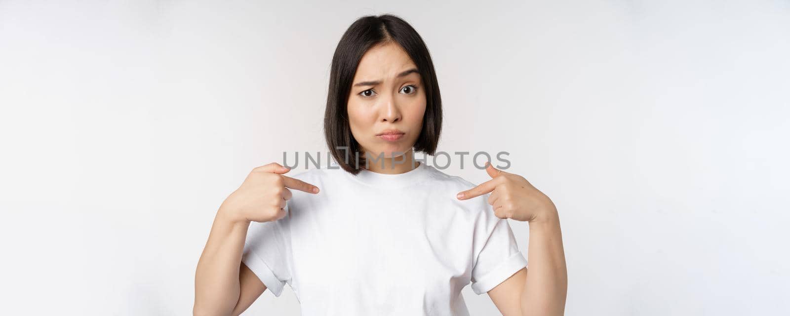Young asian woman pointing at herself with disbelief, being chosen, surprised by her candidature, standing over white background by Benzoix