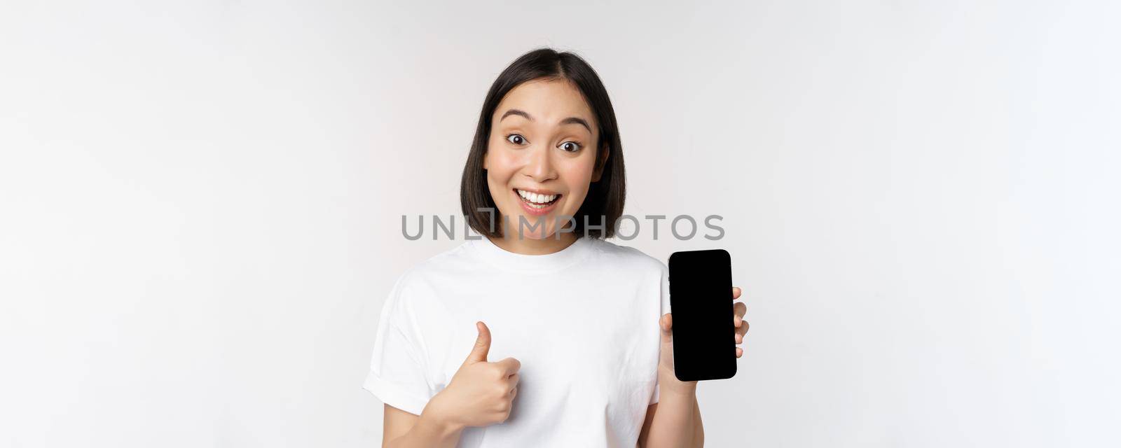 Enthusiastic young woman showing thumb up and mobile phone screen, standing in tshirt over white background by Benzoix