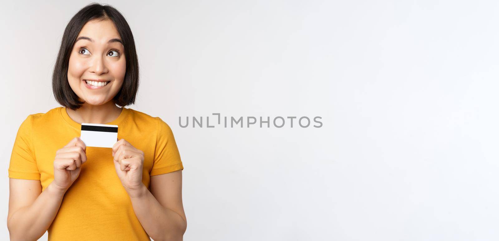 Portrait of beautiful korean girl holding credit card, recommending bank service, standing in yellow tshirt over white background by Benzoix