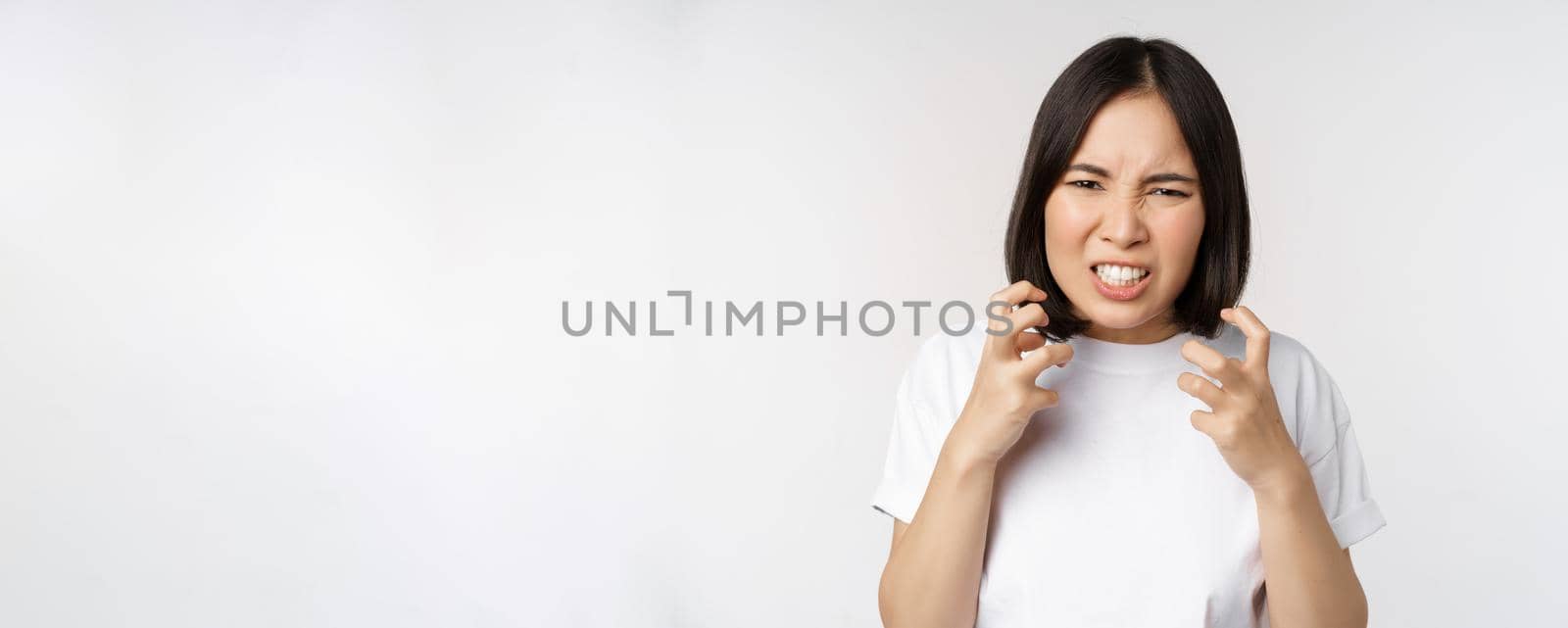 Angry asian woman cursing, looking outraged and annoyed, clench teeth and frowning furious, standing over white background.