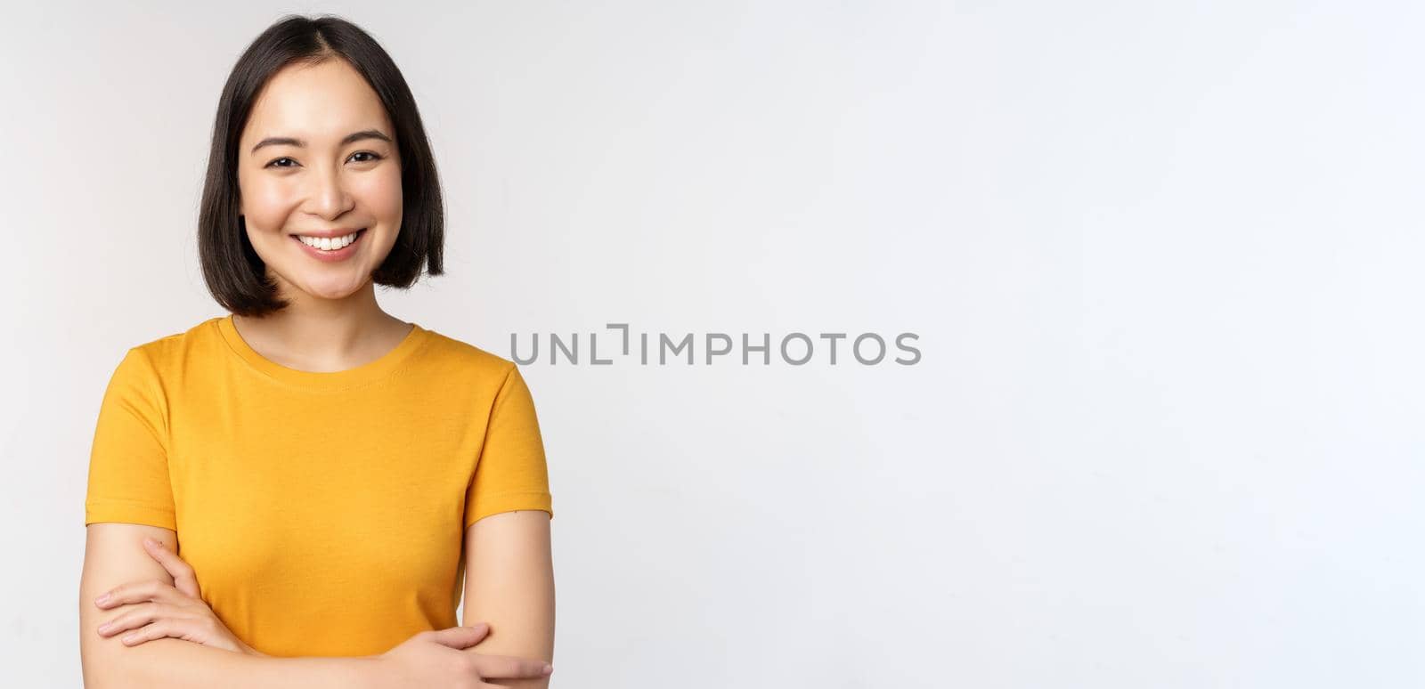 Portrait of young beautiful brunette, asian girl smiling and looking happy, cross arms on chest, standing over white background.