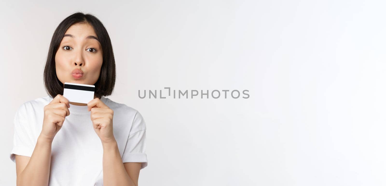 Money and finance concept. Cute japanese girl kissing her credit card, standing in tshirt over white background by Benzoix