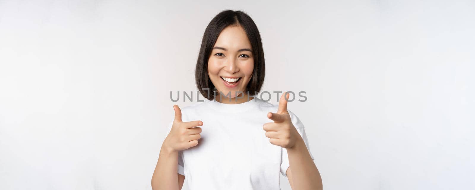 Image of smiling asian girl pointing fingers at camera, choosing, inviting you, congratulating, standing in tshirt over white background by Benzoix