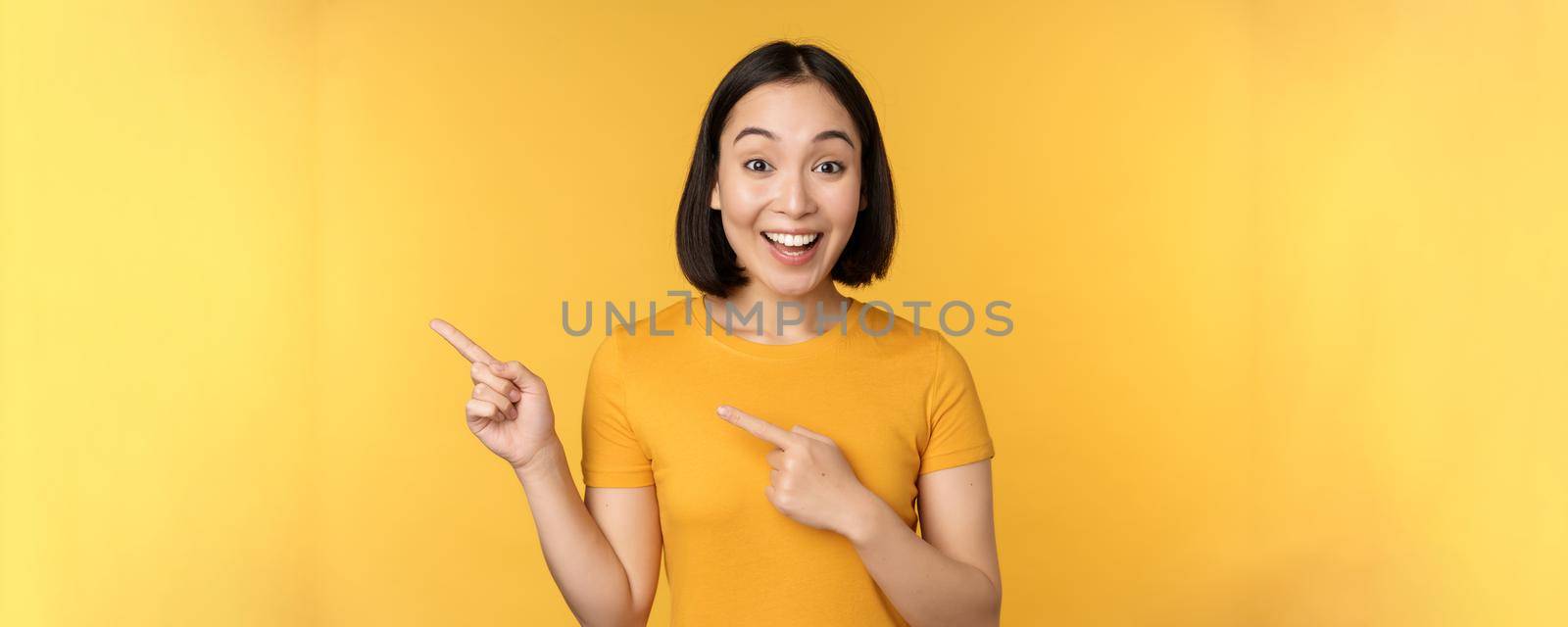 Smiling asian woman pointing fingers left, showing advertisement on empty copy space, standing over yellow background by Benzoix