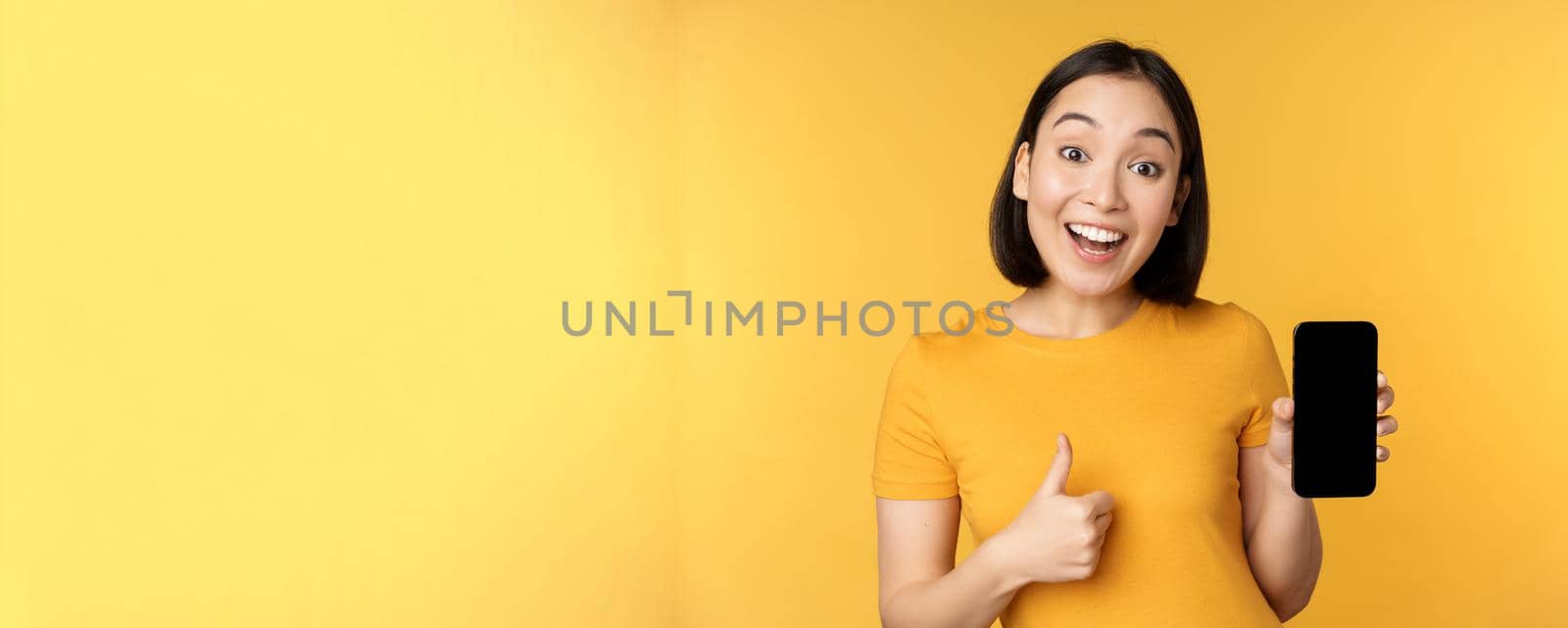 Excited asian woman showing mobile phone screen, thumbs up, like smth good, recommending smartphone application, standing over yellow background by Benzoix