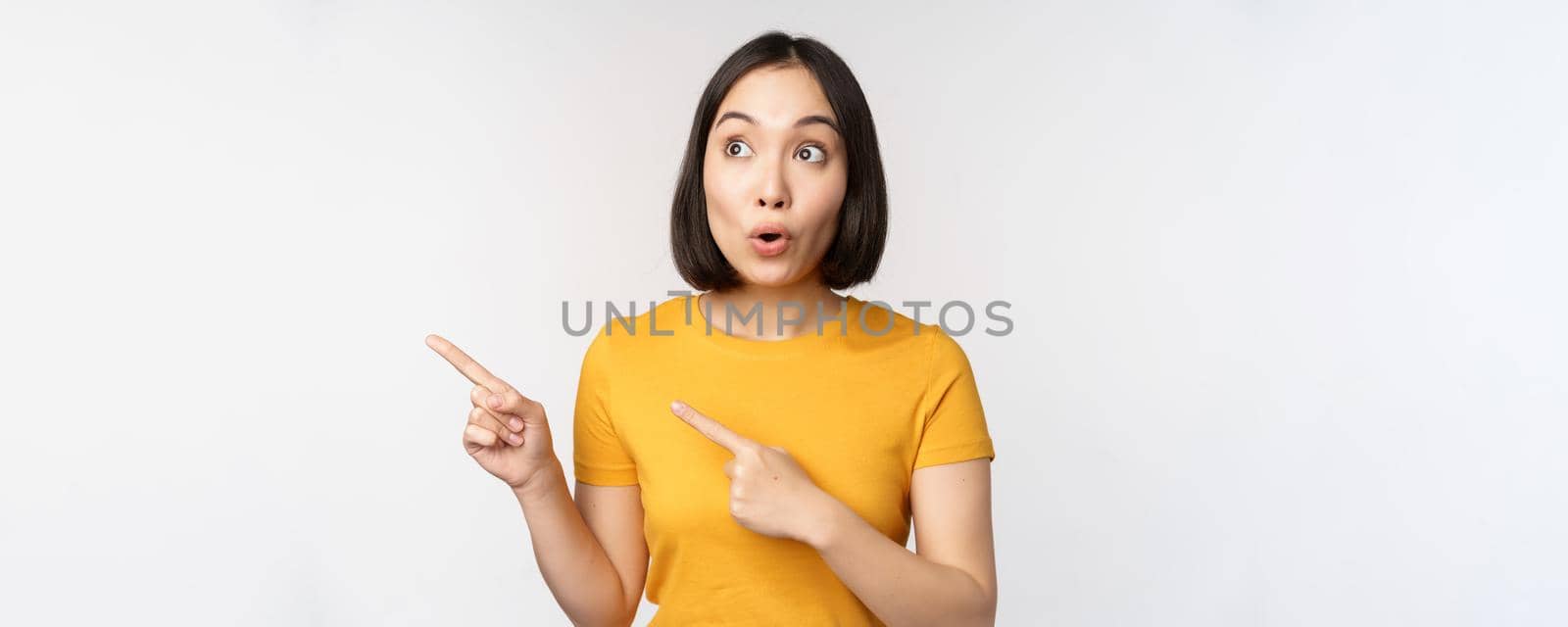 Surprised asian woman pointing and looking left at copy space, promo sale, showing advertisement with impressed face expression, standing over white backround.