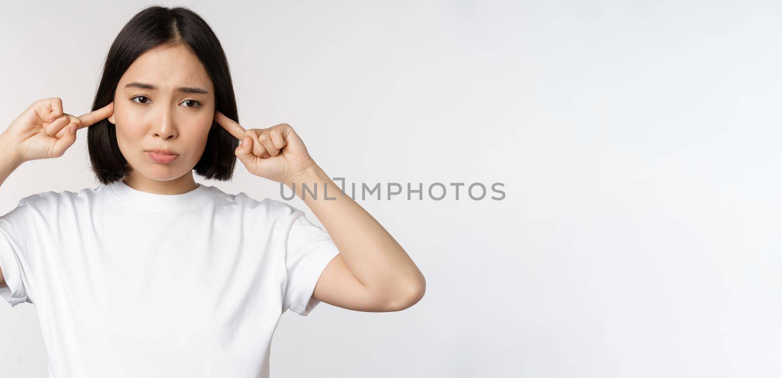 Portrait of asian woman shut ears and feeling discomfort from loud noise, annoying sound, standing over white background by Benzoix