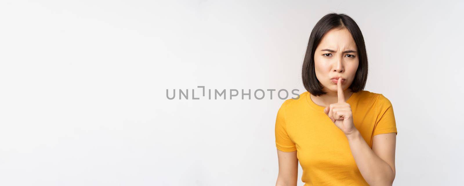 Portrait of angry asian woman shushing, press finger to lips, taboo stop talking sig, looking annoyed, scolding, standing over white background.