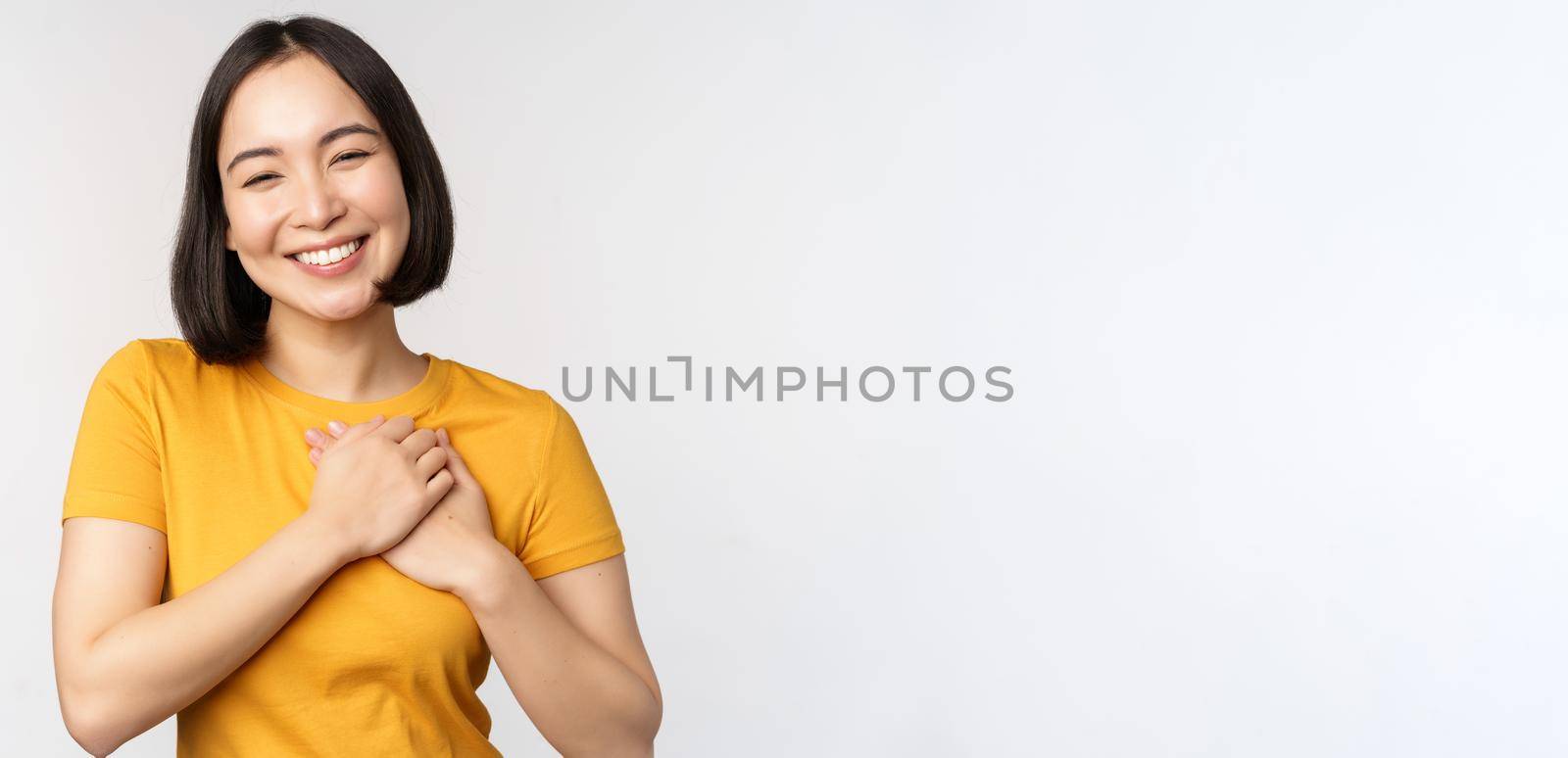 Romantic asian girfriend, holding hands on heart, smiling with care and tenderness, standing in yellow tshirt over white background.