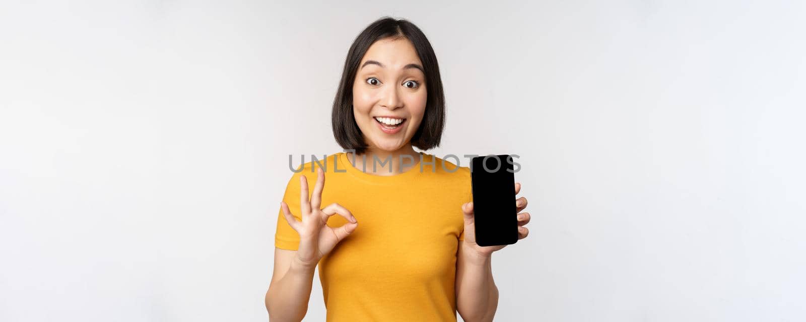 Excited asian girl showing mobile phone screen, okay sign, recommending smartphone app, standing in yellow tshirt over white background.