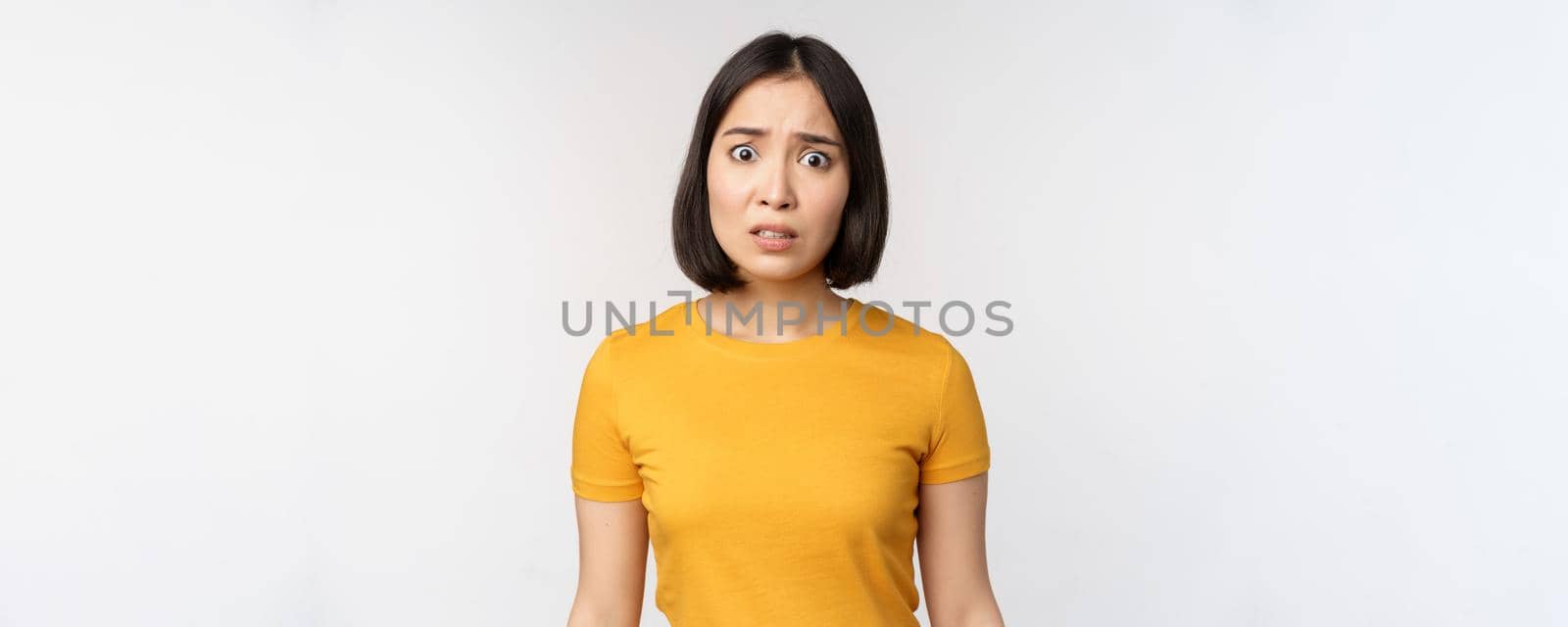 Portrait of worried korean girl, looking concerned at camera, standing in yellow tshirt over white background by Benzoix