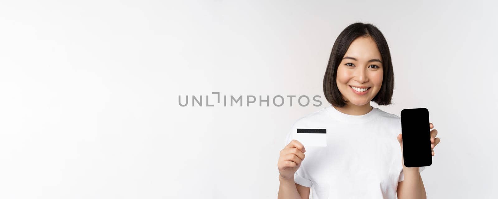 Portrait of smiling young asian woman showing mobile phone screen and credit card, standing over white background by Benzoix