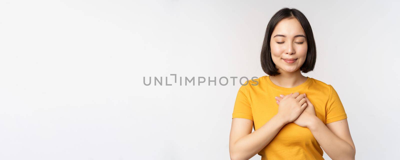 Romantic asian girfriend, holding hands on heart, smiling with care and tenderness, standing in yellow tshirt over white background.