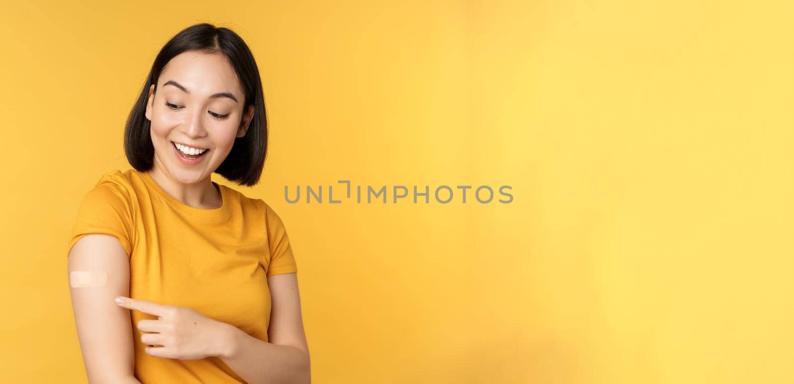 Vaccination and covid-19 pandemic concept. Happy and healthy asian girl pointing at her shoulder with band aid after vaccinating from coronavirus, yellow background.