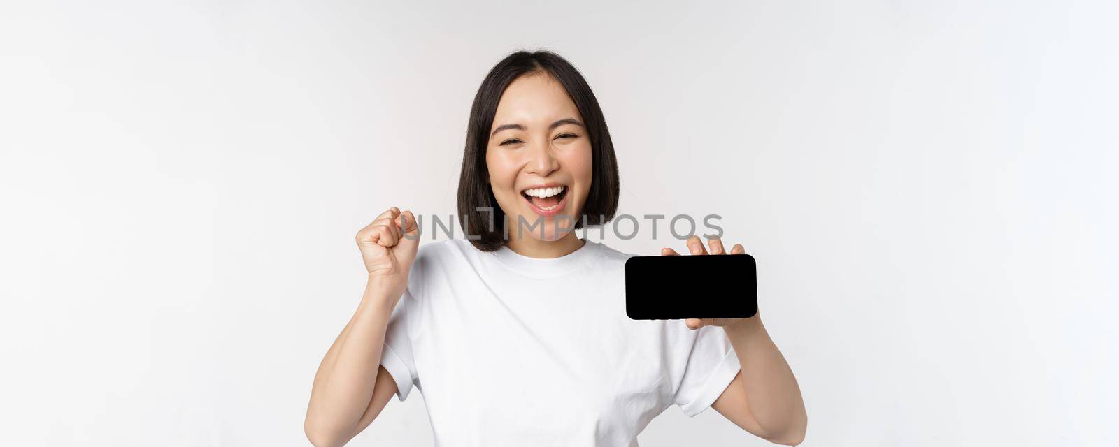 Enthusiastic asian girl scream in joy, showing horizontal smartphone screen, mobile phone display, standing over white background. Copy space