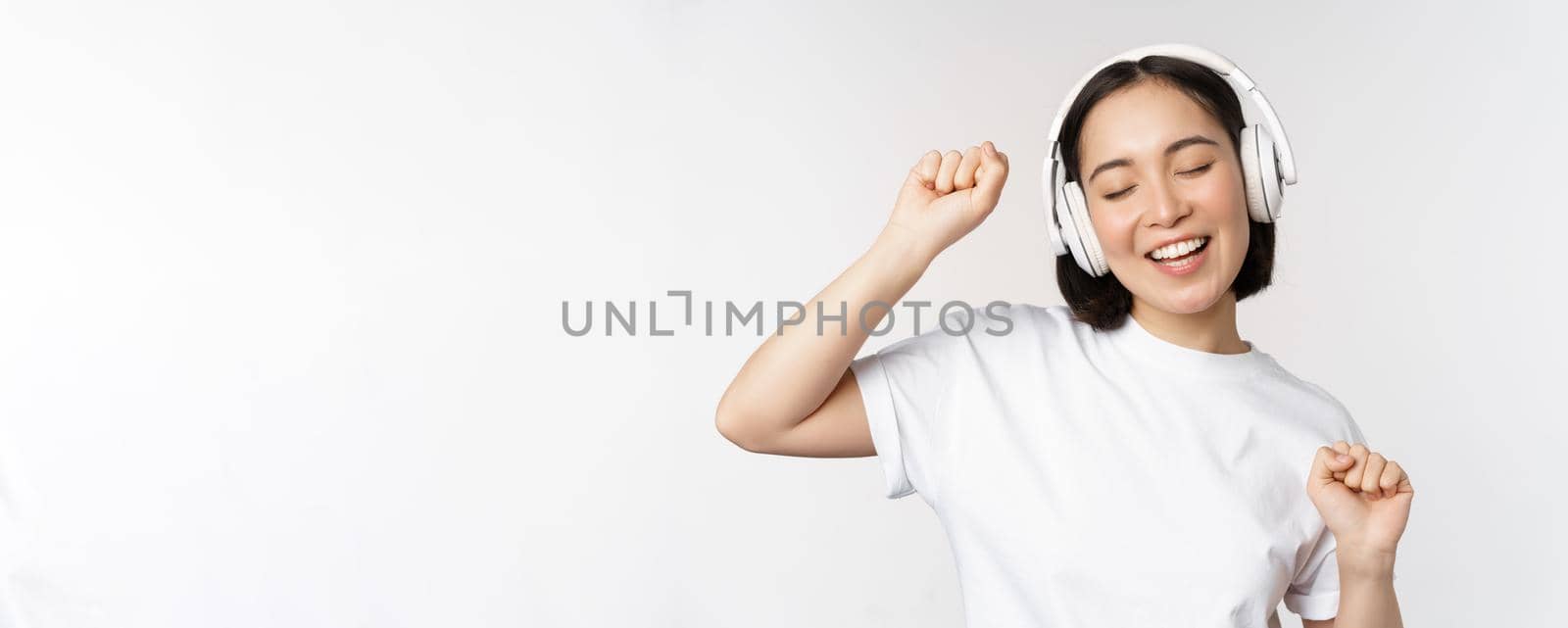 Dancing and singing asian woman, listening music in headphones, standing in earphones against white background. Copy space