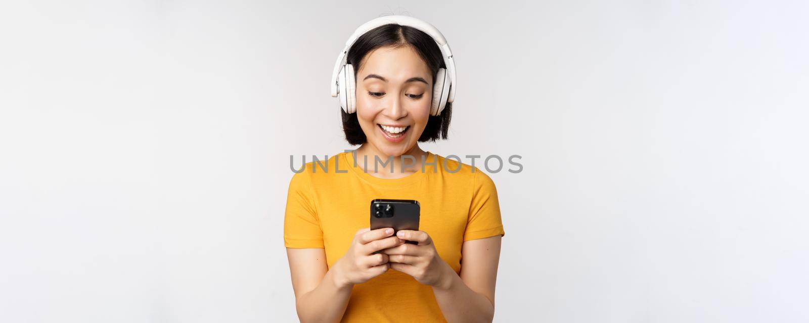 Cute japanese girl in headphones, looking at mobile phone and smiling, using music app on smartphone, standing against white background by Benzoix